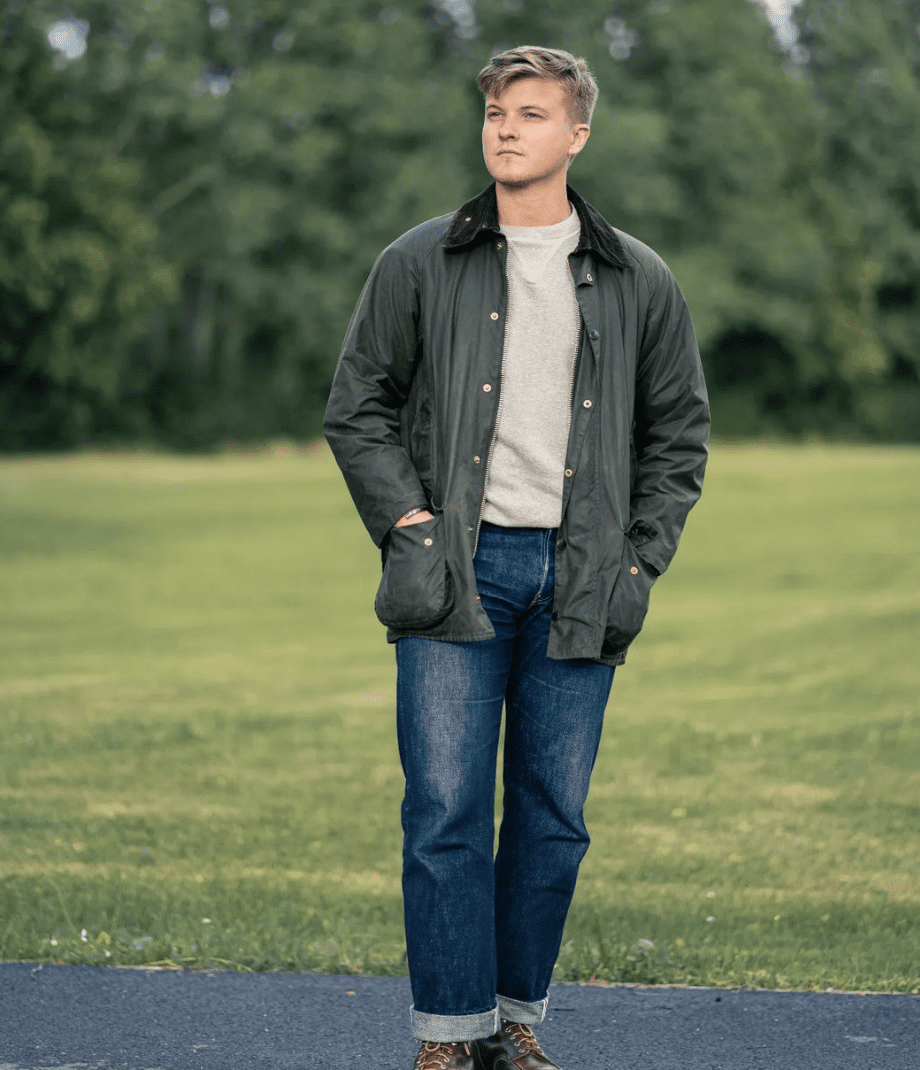 man standing in front of grass wearing a waxed jacket, blue jeans and boots