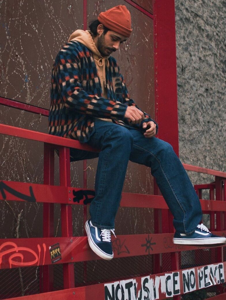 Man sitting on railing wearing sweater, colourful jacket, beanie and jeans