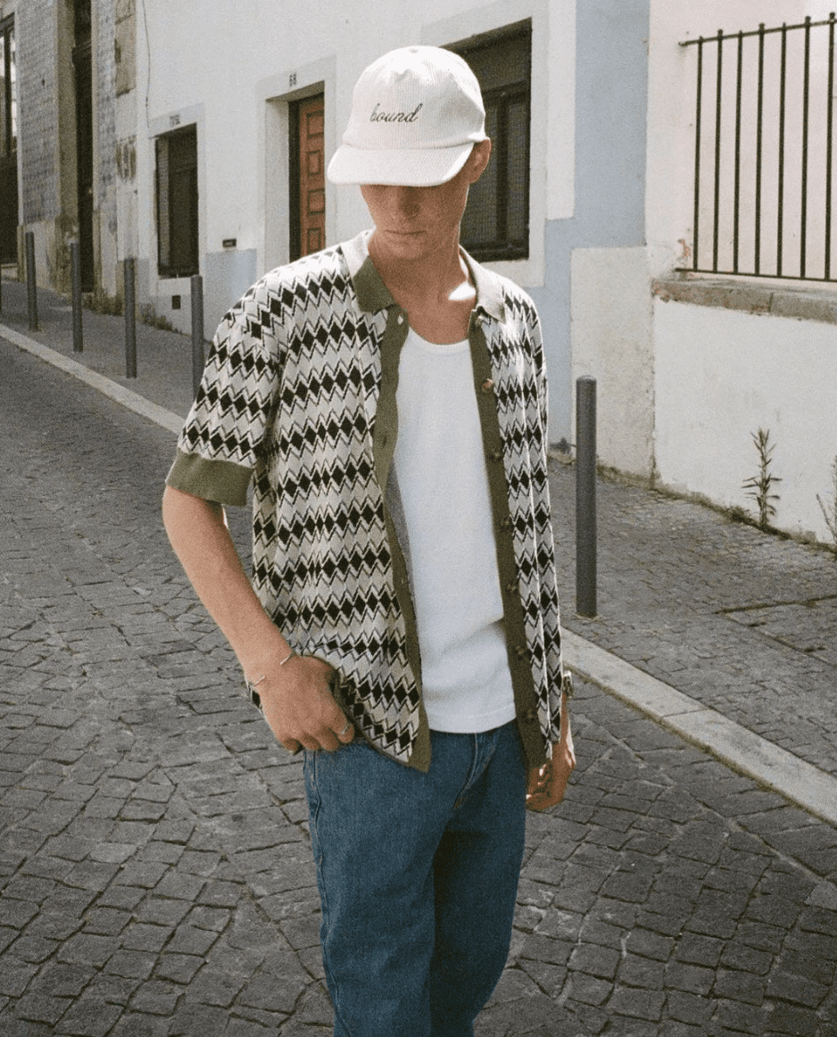 man wearing a ball cap with head down, in blue jeans standing on a street