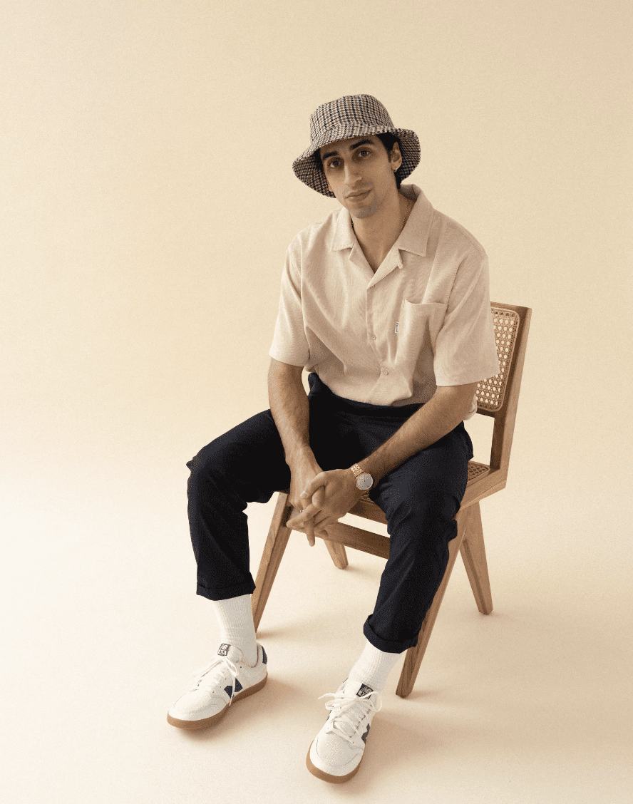 Man sitting on chair wearing a bucket hat, tshirt and jeans with a beige background