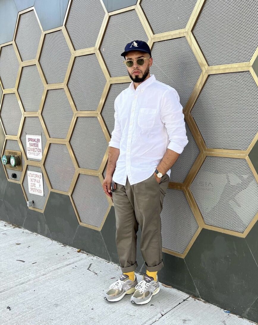 Man standing against a wall wearing sunglasses, a white button up, chinos and sneakers 