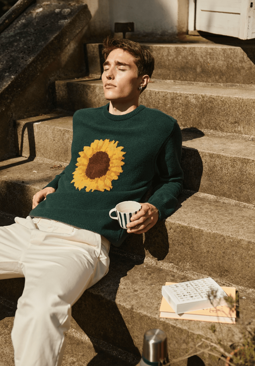 Man with eyes closed on steps wearing men's pants and a green sunflower sweater